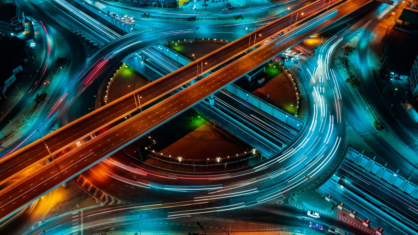 Aerial view of a brightly lit highway interchange at night, with long exposure light trails from moving vehicles creating dynamic curves and lines across multiple levels of roads.